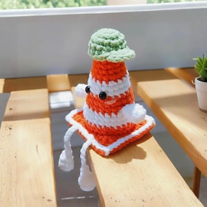 May include: A handmade crochet traffic cone toy with a green hat, white and orange stripes, and white dangling legs. The toy has black button eyes and white pom-pom arms. The cone is sitting on a wooden surface.