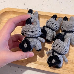 May include: Crocheted gray cat figurines with black accents, including ears, a flower, and a scarf. The handmade cat toys are displayed on a wooden tray. The cat figurines are approximately 3 inches tall.