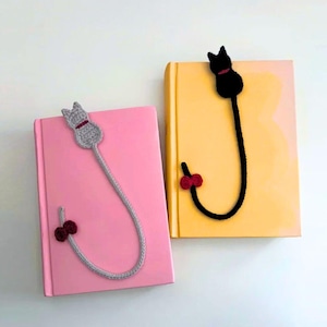 K&ouml;nnte beinhalten: Zwei geh&auml;kelte Katzen-Lesezeichen, eines grau und eines schwarz, jeweils an einem Buch befestigt. Das graue Katzen-Lesezeichen befindet sich auf einem rosa Buch, und das schwarze Katzen-Lesezeichen befindet sich auf einem gelben Buch. Beide haben eine rote Schleife.