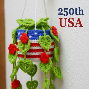 May include: A crocheted hanging plant pot featuring a red, white, and blue striped pattern with white stars on a blue background. The pot is adorned with green crocheted leaves and red flowers, suspended by white cords. Text reads 250th USA.