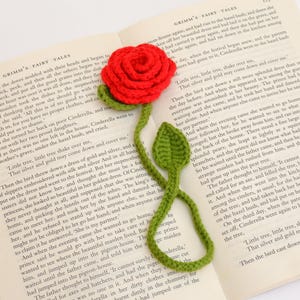 Puede incluir: Un marcapáginas de rosa roja de ganchillo con tallo y hojas verdes, colocado sobre un libro abierto. El diseño hecho a mano del marcapáginas añade un toque de elegancia a la lectura. El libro está abierto en una página de texto.