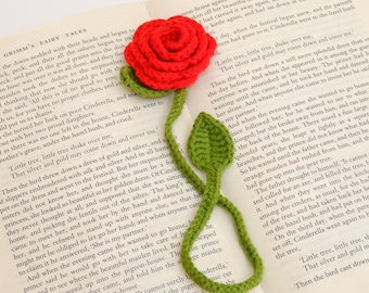 Könnte beinhalten: Ein rotes gehäkeltes Rosen-Lesezeichen mit grünem Stiel und Blättern, das auf einem offenen Buch liegt. Das handgefertigte Design des Lesezeichens verleiht dem Lesen eine elegante Note. Das Buch ist auf einer Textseite geöffnet.