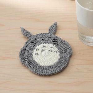 May include: A handmade crochet coaster in the shape of a cartoon character, featuring a gray outer ring and a white center with facial details. A glass of water and an open book are also on the wooden surface.