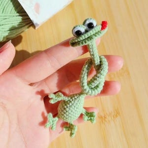 Könnte beinhalten: Ein hellgrüner gehäkelter Frosch mit Kulleraugen und roter Zunge wird in der Nähe eines Knäuels hellgrüner Wolle gehalten. Der Frosch ist geknotet und hat kleine, detaillierte Füße. Die Wolle ist mit einem weißen Etikett mit floralem Design umwickelt.
