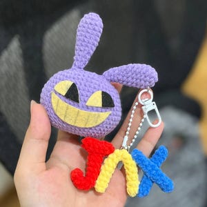 Puede incluir: Un llavero de ganchillo hecho a mano con una criatura morada con una cara sonriente y la palabra "Jinx" en rojo, amarillo y azul. El llavero tiene un cierre y una cadena plateados, perfecto para sujetar a llaves o a un bolso.