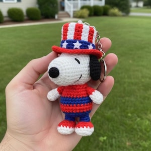 Puede incluir: Llavero de Snoopy tejido a ganchillo con sombrero y atuendo patriótico del Tío Sam. El sombrero es rojo, blanco y azul con estrellas. El atuendo es rojo y azul. El llavero se sostiene en una mano sobre un fondo verde.