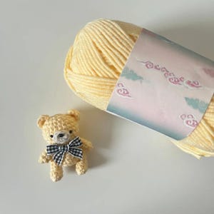 Puede incluir: Un peque&ntilde;o oso de peluche tejido a ganchillo con una pajarita a cuadros blancos y negros se encuentra junto a una madeja de lana amarilla. La lana tiene una etiqueta decorativa con un dise&ntilde;o de nubes pastel.