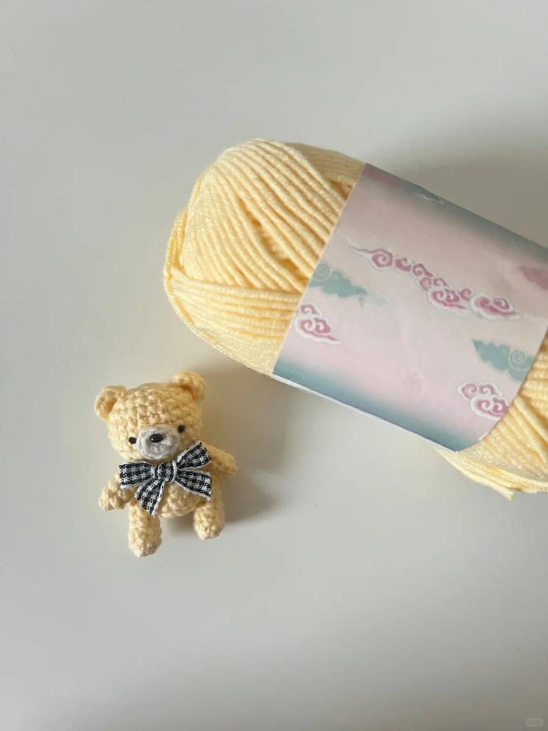 Puede incluir: Un peque&ntilde;o oso de peluche tejido a ganchillo con una pajarita a cuadros blancos y negros se encuentra junto a una madeja de lana amarilla. La lana tiene una etiqueta decorativa con un dise&ntilde;o de nubes pastel.