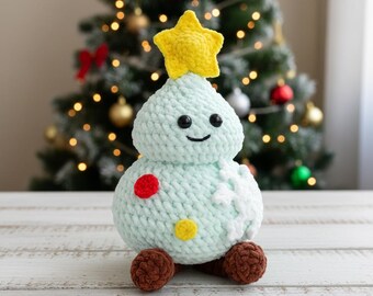 May include: A crocheted Christmas tree with a yellow star on top. The tree is light blue with a smiling face, red and yellow accents, and brown feet. The background features a decorated Christmas tree.