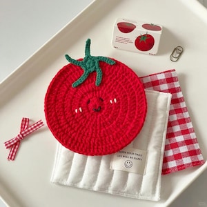 May include: A red crocheted tomato coaster with a smiling face, green stem, and white accents. A small white and red gingham bow, a white quilted cloth, and a red and white checkered napkin are also present.