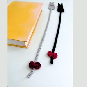 K&ouml;nnte beinhalten: Zwei handgefertigte Katzen-Lesezeichen aus H&auml;kelarbeit. Eines ist grau mit einer roten Schleife und einem Katzenkopf mit rotem Halsband. Das andere ist schwarz mit einer roten Schleife und einem Katzenkopf mit schwarzem Halsband. Ein gelbes Buch ist im Hintergrund.