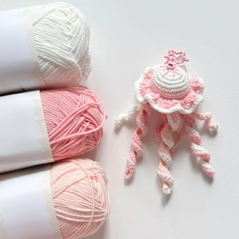 May include: A handmade crochet jellyfish in shades of pink and white, with a star-shaped embellishment. The jellyfish is next to three skeins of yarn in white, pink, and light pink. The yarn is wrapped in white paper.