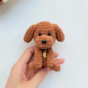 May include: A small, brown crocheted dog with floppy ears and a black nose. The dog has a black collar with a gold-colored charm. The handmade plush toy is sitting upright, showcasing its detailed stitching and soft texture.