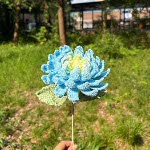May include: A handmade crochet flower in shades of light blue and yellow, with green leaves and a stem. The flower is set against a blurred background of green grass and trees.
