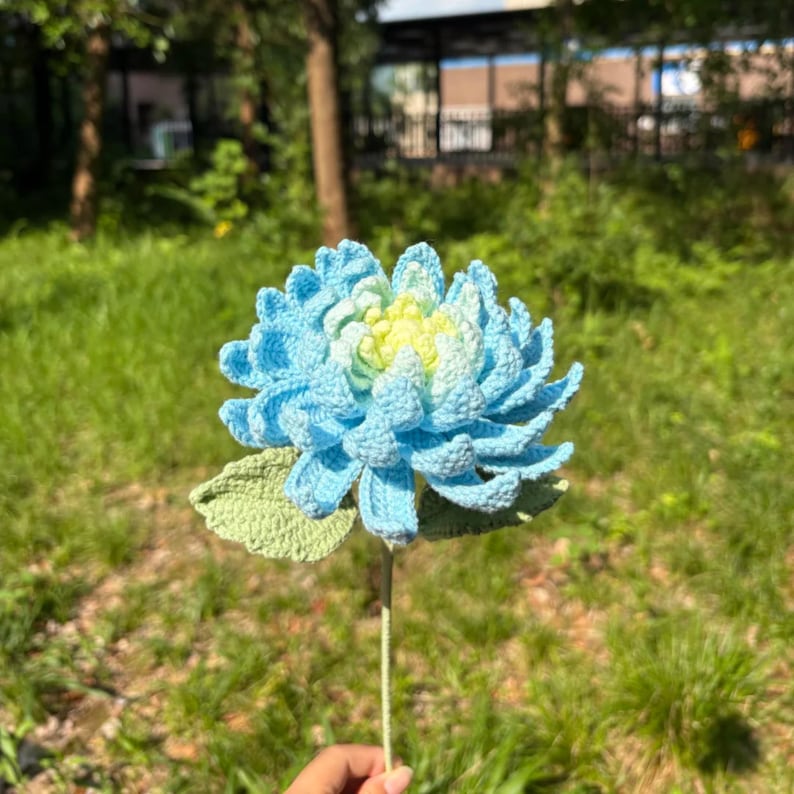 May include: A handmade crochet flower in shades of light blue and yellow, with green leaves and a stem. The flower is set against a blurred background of green grass and trees.
