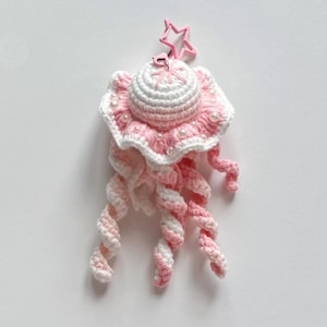 May include: A handmade crochet jellyfish keychain in pink and white. The jellyfish has a round, white top with a pink frill, adorned with faux pearls. The tentacles are a spiral of pink and white yarn, and a pink star-shaped clasp is attached.