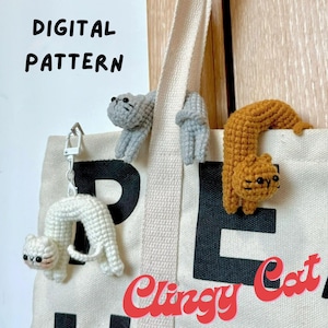 Puede incluir: Un bolso de mano de lona con las palabras "DIGITAL PATTERN" y "Clingy Cat" impresas. Varias figuras de gato de ganchillo en gris, blanco y naranja están unidas a la bolsa. Un gato blanco es un llavero.