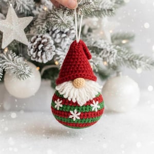 Op de afbeelding: Een handgemaakte kerstversiering in de vorm van een kabouter. De kabouter heeft een rode hoed, een witte baard en een ronde, beige neus. Het lichaam is groen en rood gestreept met witte sneeuwvlokken. De versiering hangt voor een besneeuwde kerstboom.