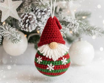 May include: A handmade Christmas ornament shaped like a gnome. The gnome has a red hat, a white beard, and a round, beige nose. The body is green and red striped with white snowflake accents. The ornament is hanging in front of a snowy Christmas tree.