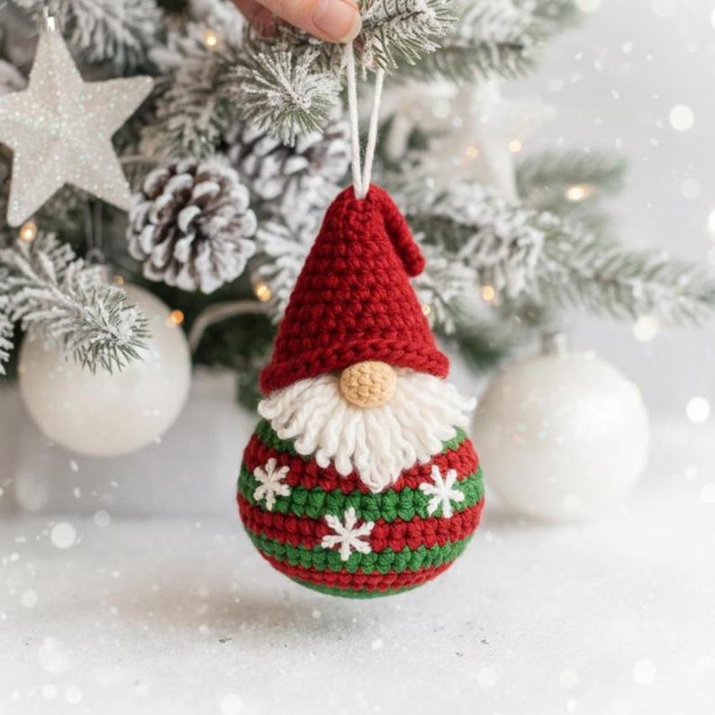 May include: A crocheted Christmas gnome ornament. The gnome features a red pointed hat, a white beard, and a beige nose. The body is striped green and red, decorated with white snowflakes. The ornament hangs in front of a snow-covered Christmas tree.