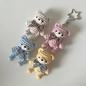 May include: Four crocheted teddy bear keychains in pastel colors: white, pink, blue, and yellow. Each bear has a black and white checkered bow and black button eyes. A silver star-shaped charm is attached to one of the bears.