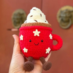 Op de afbeelding: Een rode pluche koffiekop met witte steraccenten en een lachend gezicht. De kop is bedekt met een witte slagroom met bruine spikkels. Het heeft bruine benen en een rood handvat.