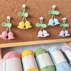 May include: Crocheted cherry-shaped ornaments in pink, yellow, light blue, and lavender, with green leaves and white pearl accents. The ornaments are displayed on a corkboard. Below are skeins of yarn in matching colors.