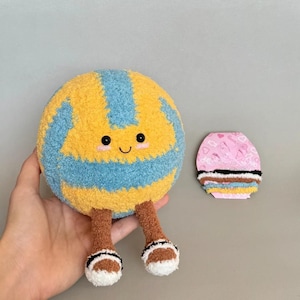 K&ouml;nnte beinhalten: Ein Pl&uuml;sch-Volleyball-Spielzeug mit einem l&auml;chelnden Gesicht, gelben und blauen Paneelen und braunen Beinen mit wei&szlig;en und schwarzen Akzenten. Ein kleines rosa und wei&szlig;es Dekorationsobjekt befindet sich neben dem Volleyball.
