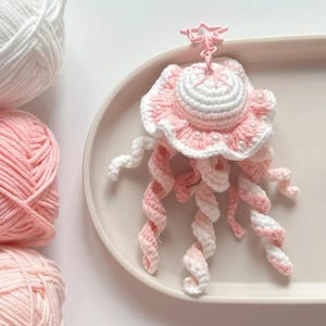 May include: A handmade crochet jellyfish in shades of pink and white, adorned with small pearl beads. The jellyfish has a star-shaped clasp and long, spiraling tentacles. The craft project is displayed on a light pink tray, with balls of yarn in the background.