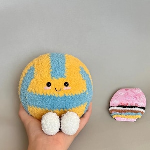 K&ouml;nnte beinhalten: Ein gelb-blauer Pl&uuml;sch-Volleyball mit einem l&auml;chelnden Gesicht und wei&szlig;en Pompon-F&uuml;&szlig;en. Rechts vom Volleyball befindet sich ein rosa-braunes Dekorationsobjekt.