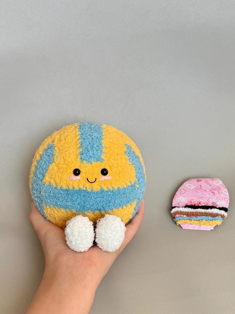 K&ouml;nnte beinhalten: Ein gelb-blauer Pl&uuml;sch-Volleyball mit einem l&auml;chelnden Gesicht und wei&szlig;en Pompon-F&uuml;&szlig;en. Rechts vom Volleyball befindet sich ein rosa-braunes Dekorationsobjekt.