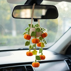May include: A crocheted hanging ornament featuring a small beige basket containing several bright orange pumpkins with green vines, leaves, and small yellow flowers, suspended by a beige cord from a car rearview mirror.