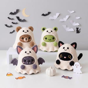 May include: Four crocheted plush toys dressed in white ghost costumes. The figures include a brown bear, a green frog, a purple bat with wings, and a black cat with ears and a tail. Each ghost hood features a stitched smiling face.