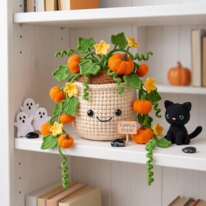 May include: A crocheted cream-colored pot with a smiling face, filled with green vines, yellow flowers, and orange pumpkins. A small wooden sign reads PUMPKIN PICKIN'. Beside the pot sits a small black crocheted cat and white ghost cutouts.
