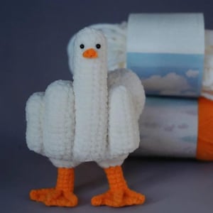 Op de afbeelding: Een handgemaakte witte gehaakte gans met oranje poten en snavel, die de middelvinger opsteekt. De gans staat voor twee bollen wol. De achtergrond is effen grijs.