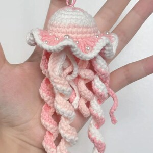 May include: A handmade crochet jellyfish keychain in shades of pink and white. The jellyfish has a rounded top with a ruffled edge, adorned with small white beads. The tentacles are long and spiraled. The keychain hardware is pink.