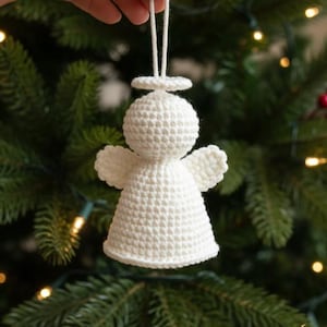 以下が含まれることがあります： 白いかぎ針編みの天使のクリスマスオーナメント。ハローと翼が付いています。天使は白い紐で吊り下げられており、クリスマスツリーに飾る準備ができています。背景は、緑のクリスマスツリーのぼやけたビューです。