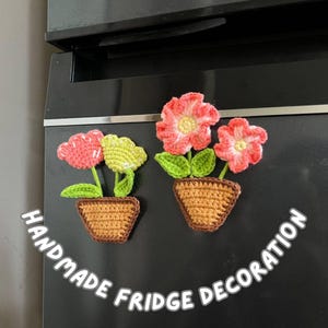 May include: Handmade crochet fridge decoration showing two flower pot designs. One pot has a pink and yellow flower, the other has two pink flowers. The pots are brown, with the text "HANDMADE FRIDGE DECORATION" visible.