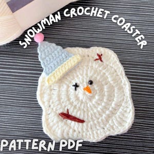 Peut inclure: Un dessous de verre fait à la main en crochet blanc représentant un bonhomme de neige avec un chapeau bleu, un bec orange et des yeux noirs. Le dessous de verre a un design circulaire et le texte "SNOWMAN CROCHET COASTER" et "PATTERN PDF" sont visibles.