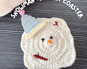 May include: A handmade, white crochet snowman coaster with a blue hat, orange beak, and black eyes. The coaster has a circular design and the text "SNOWMAN CROCHET COASTER" and "PATTERN PDF" are visible.