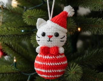 May include: A handmade crochet Christmas ornament shaped like a white cat wearing a red Santa hat and bow tie. The cat's body is a red and white striped ball. The ornament hangs from a white string and is set against a green Christmas tree.