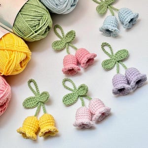 Puede incluir: Flores en forma de campana hechas a ganchillo en varios colores, incluyendo rosa, amarillo, azul y morado, con hojas verdes. Las flores hechas a mano están dispuestas sobre una superficie blanca, con ovillos de lana en el fondo.