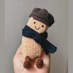 May include: A plush, beige-colored toy with a smiling face, wearing a dark gray cap and a navy blue scarf. The toy has brown feet and is designed to look like a character. The toy is handmade.