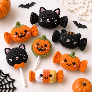 Könnte beinhalten: Eine Sammlung gehäkelter Halloween-Artikel, darunter schwarze Katzenfiguren mit Flügeln, orangefarbene kürbisförmige Bonbons, ein runder Kürbis-Lutscher, ein schwarzer Katzen-Lutscher mit Fliege und eine weiß-orange-gelbe Zuckermais-Figur.