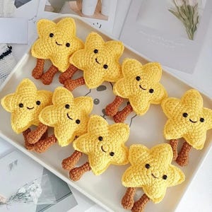 Puede incluir: Una colección de juguetes en forma de estrella, tejidos en amarillo, con caras sonrientes y piernas marrones, dispuestos en una bandeja blanca. Los juguetes hechos a mano tienen un diseño caprichoso, ideal para niños.