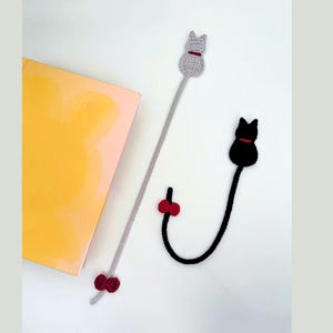 K&ouml;nnte beinhalten: Zwei handgefertigte Katzen-Lesezeichen aus H&auml;kelarbeit. Eines ist grau mit rotem Kragen und Schleife, das andere ist schwarz mit rotem Kragen. Die Lesezeichen liegen neben einem gelben Buch.