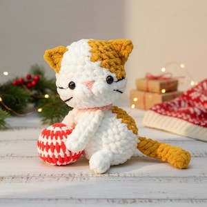 May include: A handmade crochet cat doll in white and orange, holding a red and white ball. The cat has black eyes, a pink nose, and a pink collar. The tail and ears are orange. The doll is sitting on a white wooden surface.