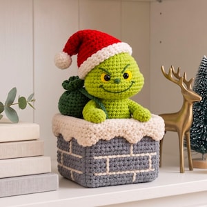 May include: A crocheted green creature with yellow eyes and a mischievous smile, wearing a red and white Santa hat. It sits inside a gray crocheted chimney with white trim, holding a dark green sack over its shoulder, crafted from yarn.