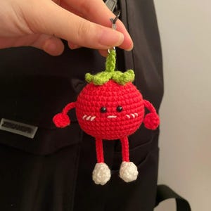 Puede incluir: Un llavero de fresa roja de ganchillo con un tallo y hojas verdes. La fresa tiene una cara linda con brazos y piernas pequeños, y detalles blancos en los pies. El llavero está sujeto a un anillo plateado.