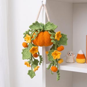 Könnte beinhalten: Eine gehäkelte Hängedekoration mit einem zentralen orangefarbenen Kürbis, umgeben von grünen Blättern, geschwungenen Ranken und gelben Blüten. Das Objekt ist an einem dicken, cremefarbenen, gedrehten Seil befestigt und wirkt handgefertigt.